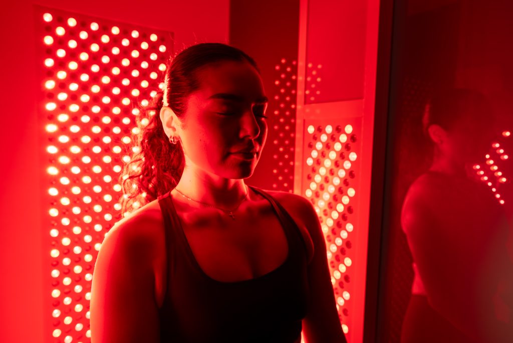 A woman with closed eyes stands in front of an illuminated red light therapy panel in a dimly lit room.