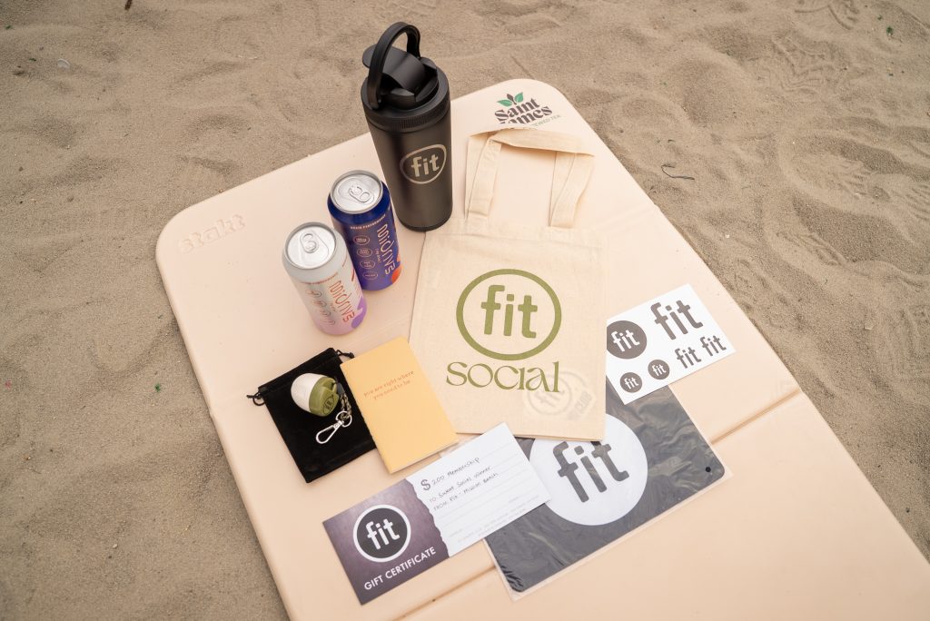 A sand-colored mat on the beach holds Fit Social merchandise, including a tote bag, drinkware, canned drinks, stickers, a gift certificate, a keychain, and a small booklet.