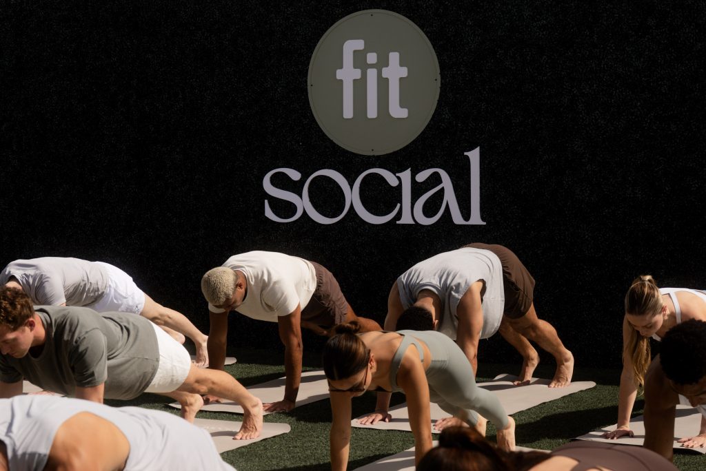 A group of people in athletic wear perform a plank exercise on mats outdoors in front of a wall with the words “fit social” displayed.