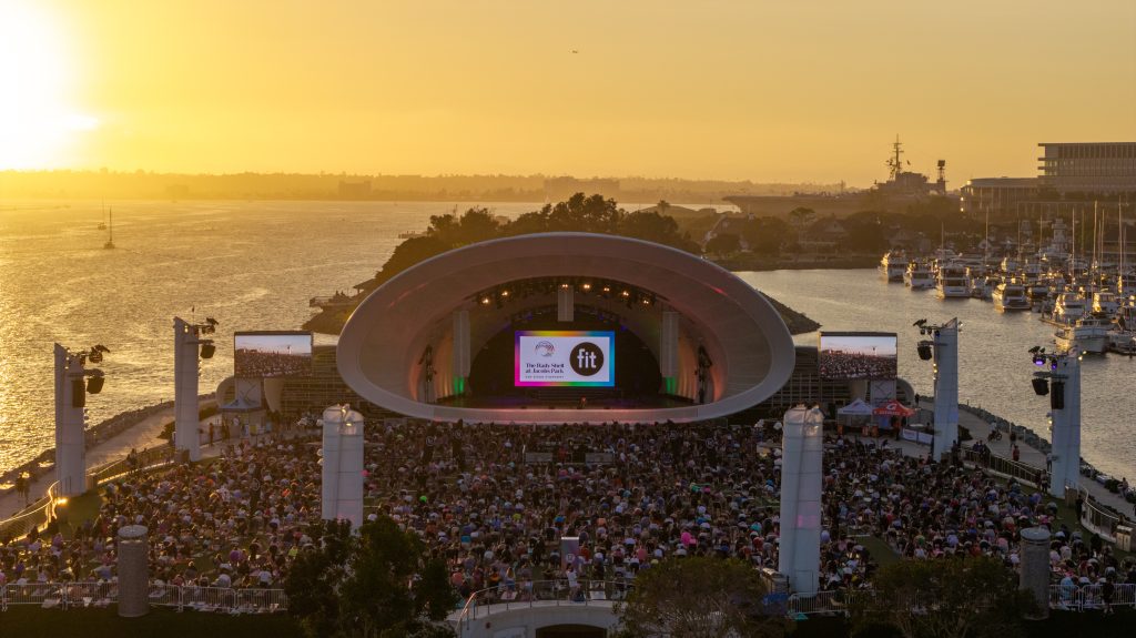Outdoor amphitheater at sunset with a large audience, stage, and water with boats in the background; screens display logos.