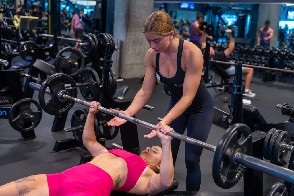 A woman in pink workout attire bench presses a barbell while another woman in black gym clothes spots her in a gym with various equipment.